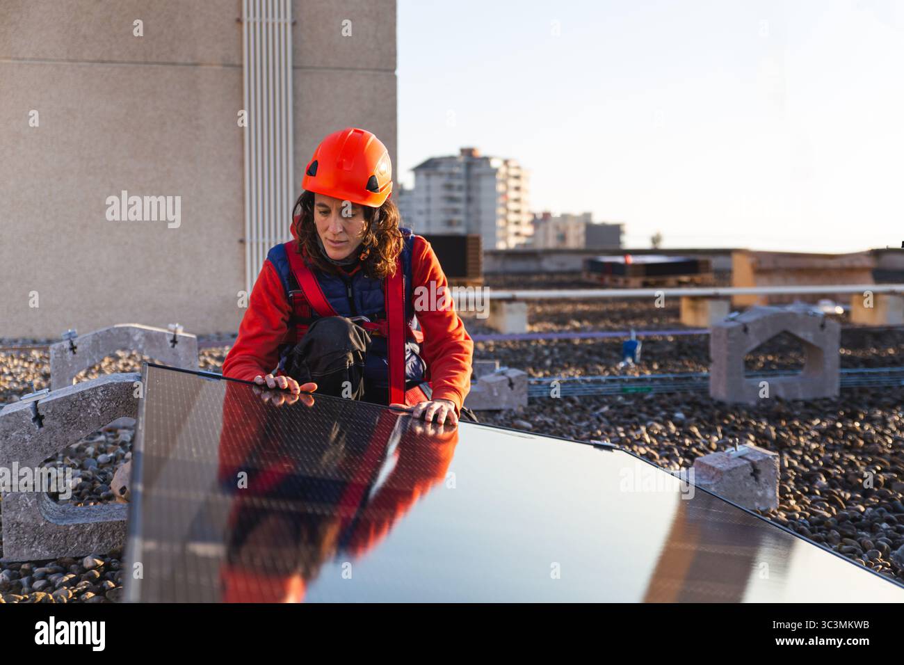 Ein Arbeiter in einem Schutzhelm inspiziert ein Solarpanel während der Installation auf einem gewerblichen Dach. Der städtische Hintergrund unterstreicht den Wandel der Stadt zur Erneuerung Stockfoto
