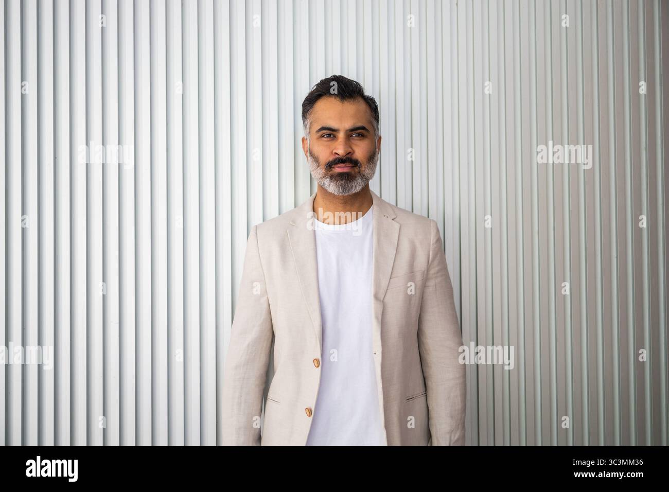 Ein selbstbewusster Indianer posiert vor einem gestreiften Hintergrund, trägt einen beigefarbenen Blazer über einem weißen T-Shirt und verkörpert Sommerstyle und mühelose Sophisti Stockfoto