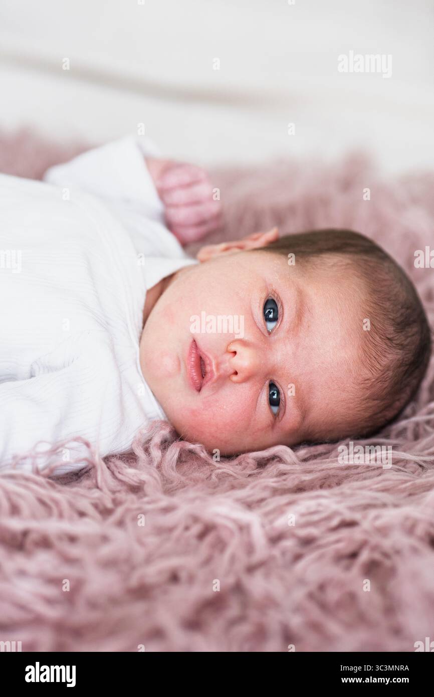 Ein neugeborenes Baby, das friedlich auf einem weichen rosa Teppich liegt, in einen gemütlichen weißen Einteiler gewickelt. Die sanfte Textur und das ruhige Ambiente schaffen ein warmes, behagliches A Stockfoto