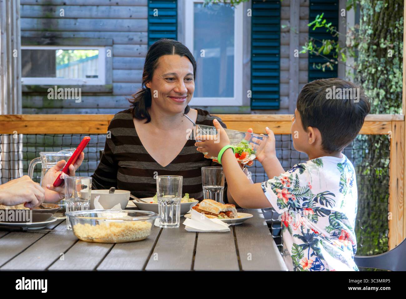 Eine Familie teilt sich eine köstliche Mahlzeit auf der Holzterrasse einer Ökotourismushütte, die von Wald umgeben ist. Gemeinsam erleben sie Natur und Entspannung in A Stockfoto
