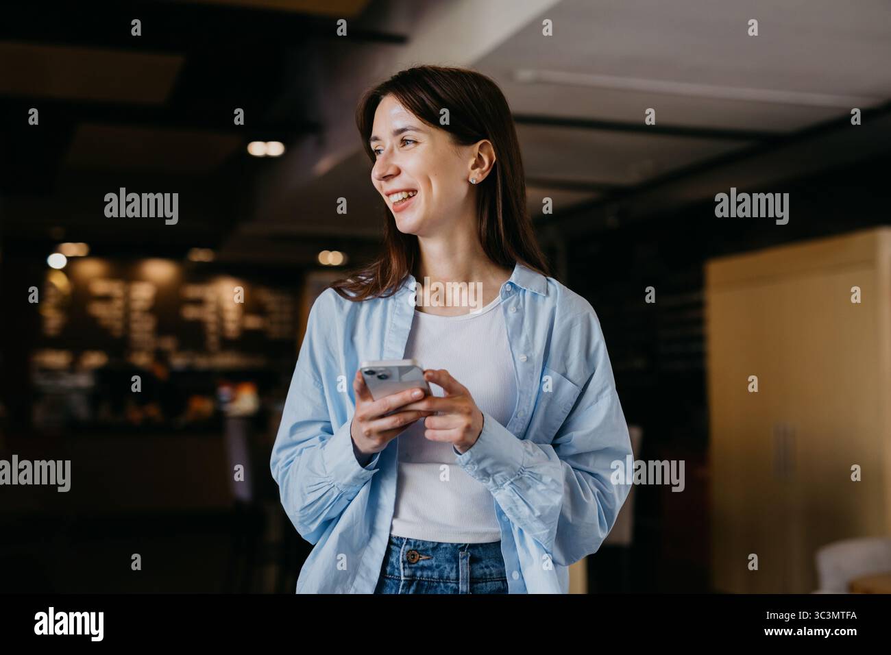 Eine Frau in einem lässigen blauen Hemd und weißem Oberteil steht lächelnd und hält ein Smartphone. Sie arbeitet aus der Ferne in einem gemütlichen Café Stockfoto