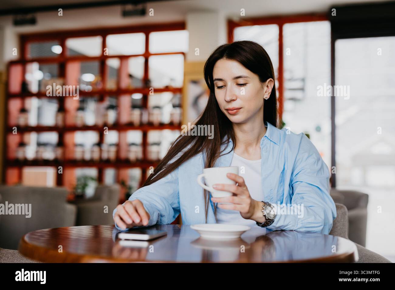 Eine Frau sitzt in einem gemütlichen Café und genießt eine Tasse Kaffee, während sie an ihrem Smartphone arbeitet. Das entspannte Ambiente schafft die perfekte Arbeitsumgebung für Stockfoto