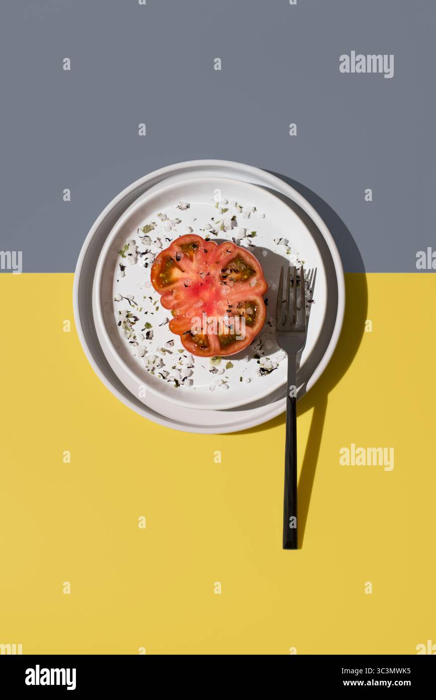 Blick von oben auf eine in zwei Hälften geschnittene raf-Tomate, die mit Salz und Gewürzen bestreut und elegant auf einem weißen Teller mit Gabel platziert ist. Der lebhafte gelbe und schwule Backgrou Stockfoto