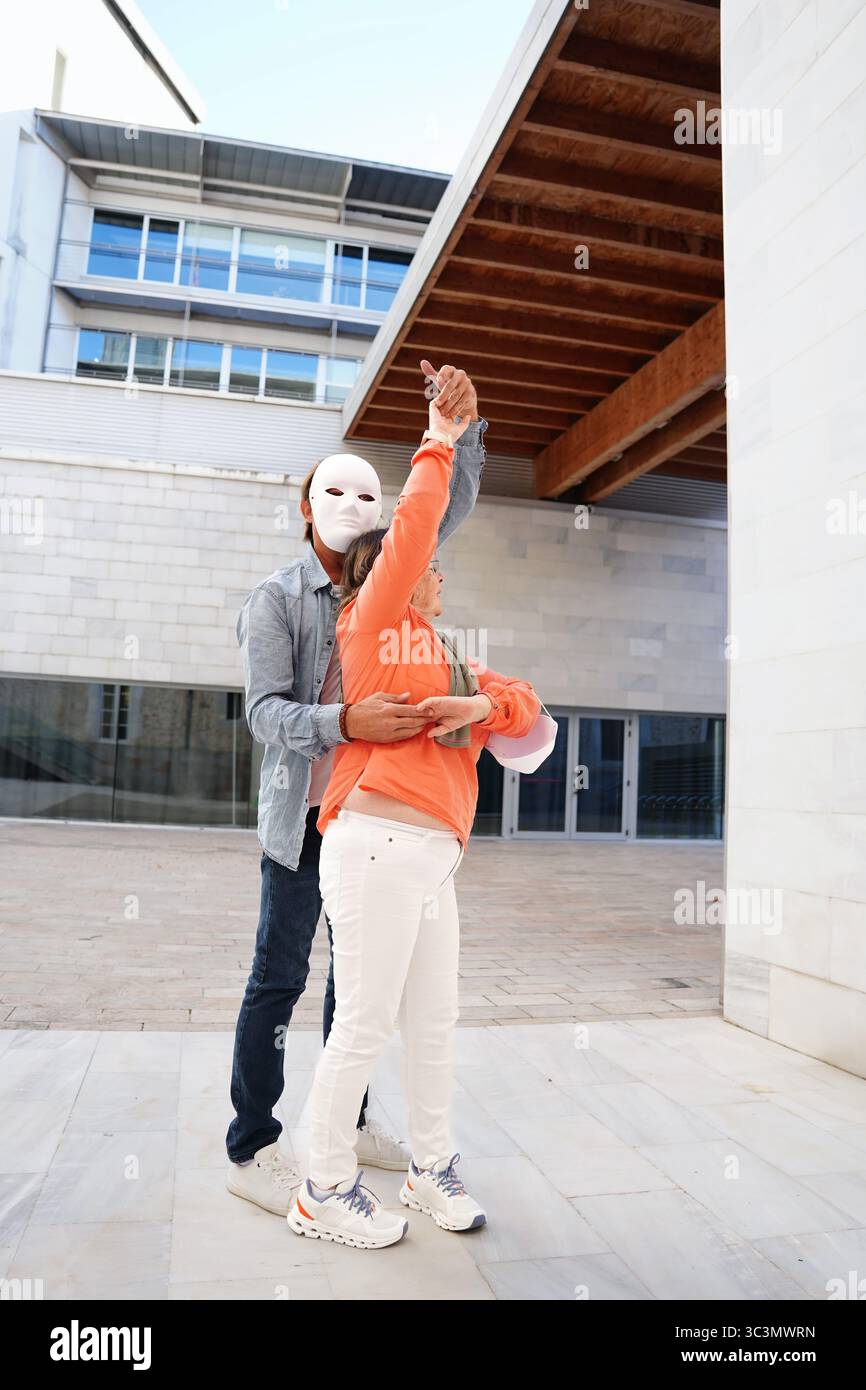 Zwei Personen, einer trägt eine weiße Maske, nehmen an einer Aktivität im Freien in einer modernen architektonischen Umgebung Teil. Ihre synchronisierten Bewegungen erzeugen Stockfoto