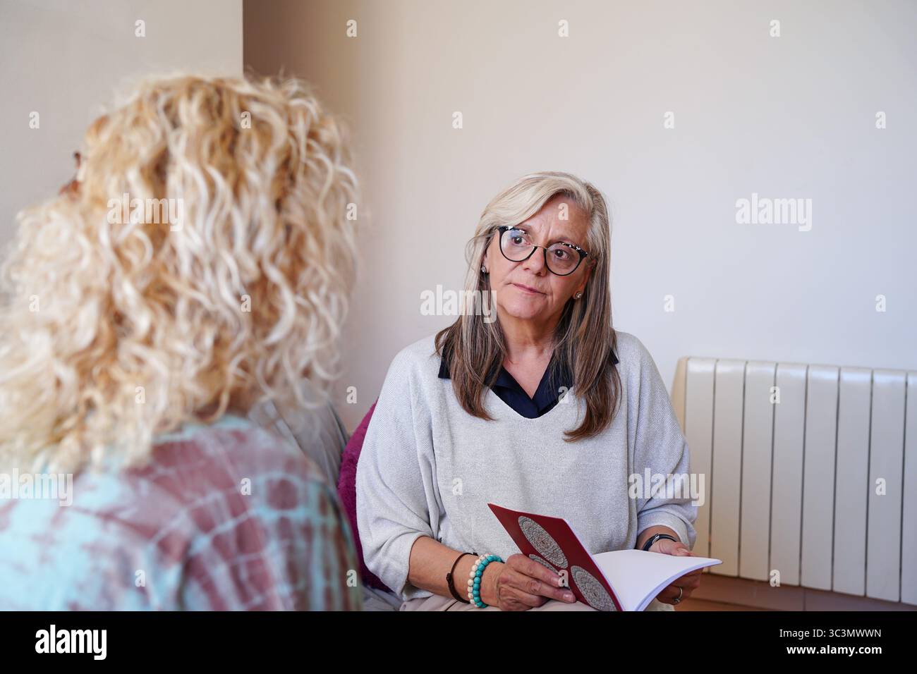 Eine ältere Frau hört ihrem Freund aufmerksam zu, während sie ein Buch hält, und hebt die Wärme und die Verbindung hervor, die sie in einem gemütlichen Indoor-Set teilen Stockfoto