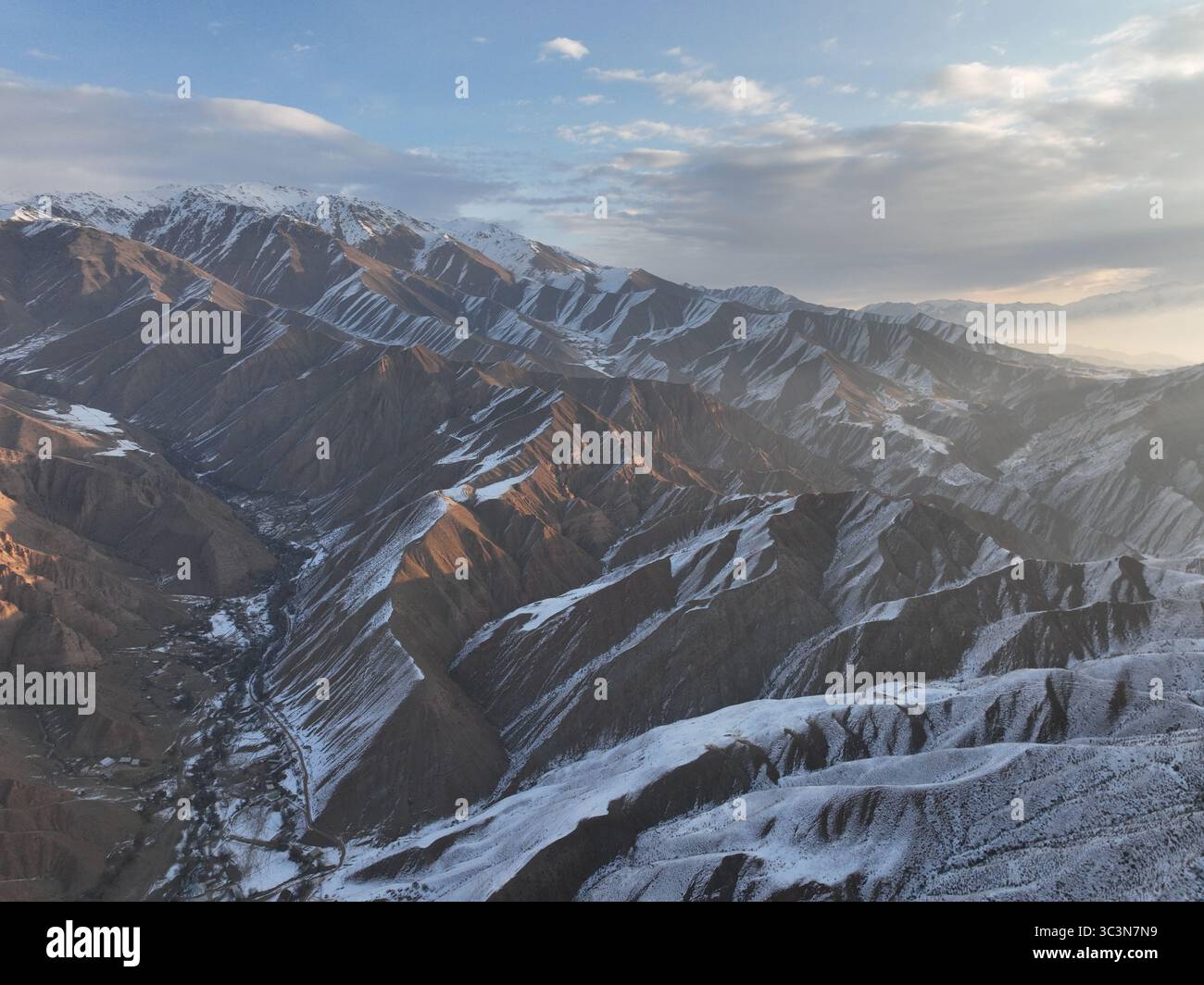 Ein weites Luftbild, das komplexe Berglandschaft mit tiefen Tälern, beträchtlicher Schneedecke und einem fernen Dorf unter weichem, diffusem Licht enthüllt Stockfoto