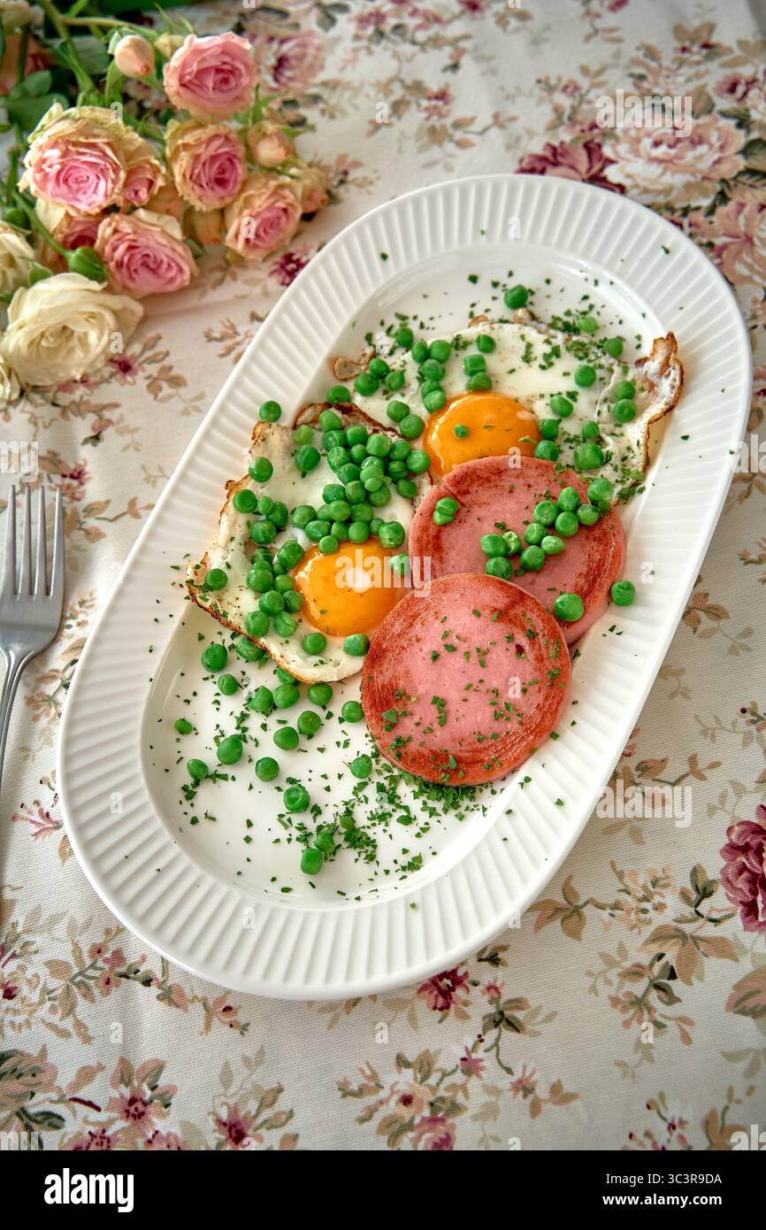 Ein gesunder Frühstücksteller mit Eiern, Schinken und frischen grünen Erbsen auf einer Tischdecke mit Blumenmuster für einen angenehmen Start in den Morgen Stockfoto