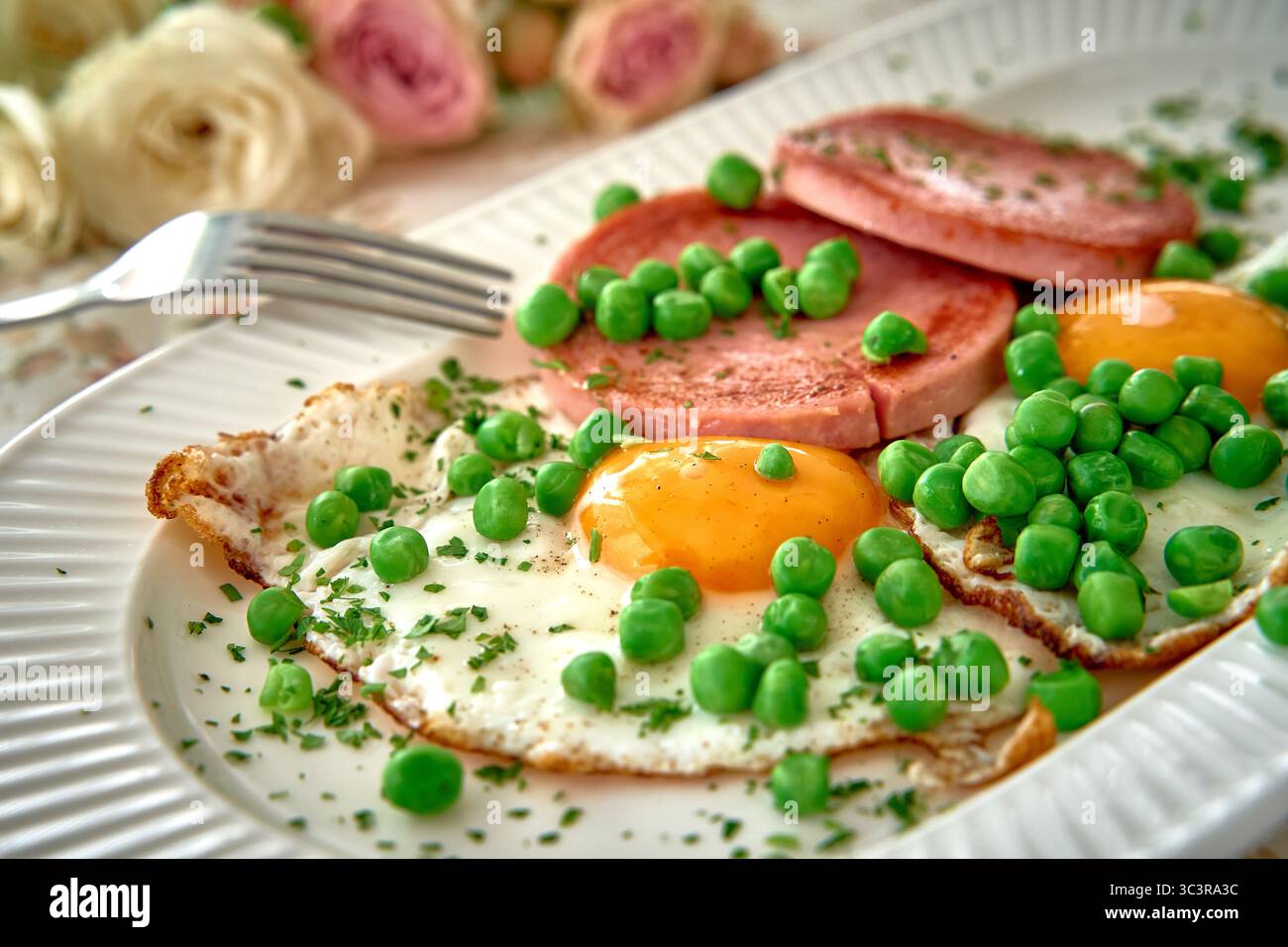 Köstlicher Frühstücksteller mit sonnigen Eiern, geschnittenem Schinken und frischen grünen Erbsen, die auf einem Deko-Teller liegen und von wunderschöner Blüte umgeben sind Stockfoto