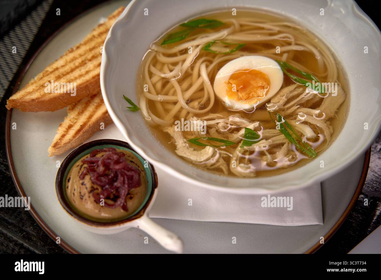 Eine Schüssel warmer und wohltuender Nudelsuppe mit zarten Nudeln, einem weich gekochten Ei und herzhafter Brühe, begleitet von geröstetem Brot und einem schmackhaften Geschmack Stockfoto