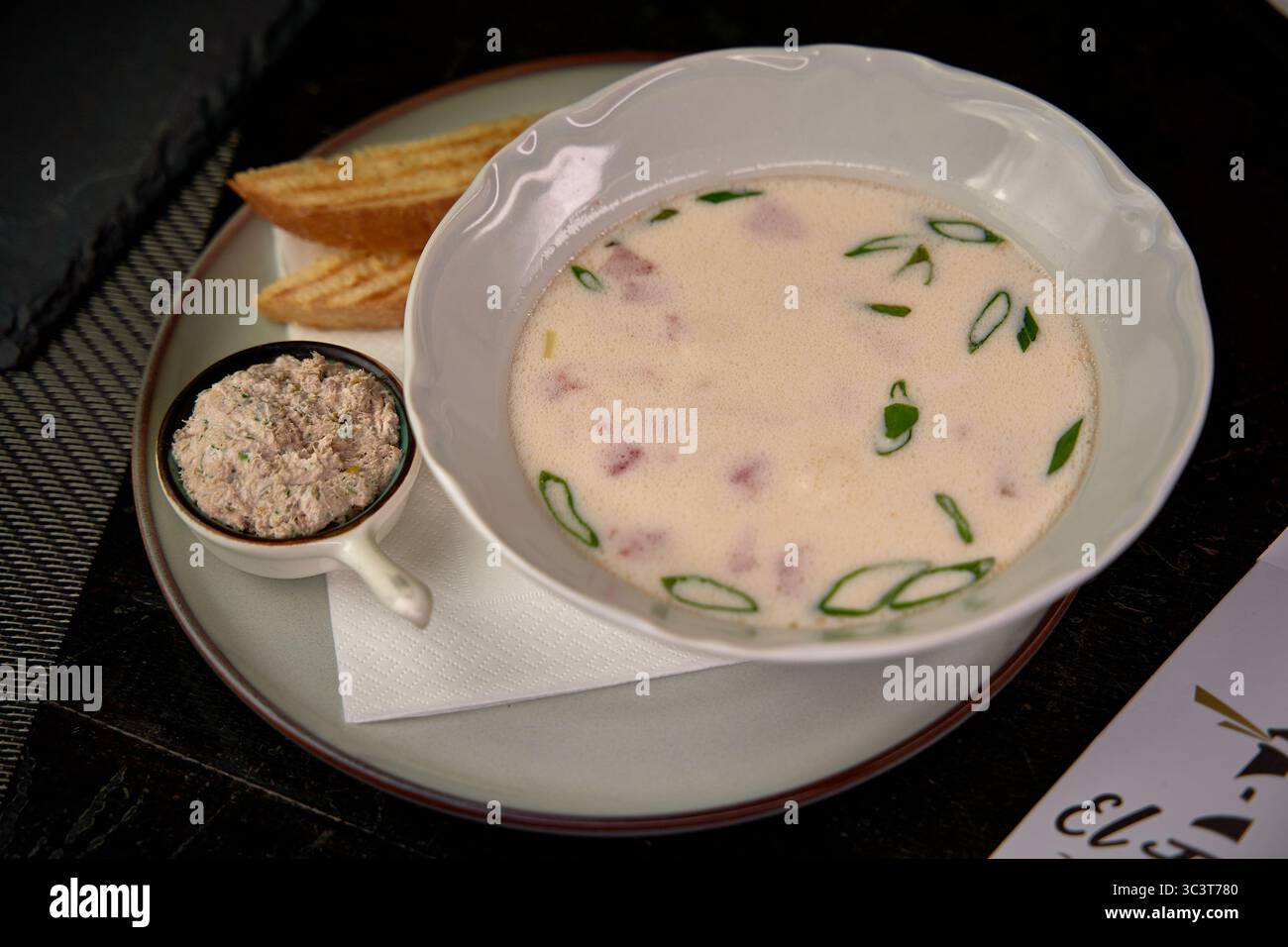 Eine Schüssel cremige Suppe garniert mit grünen Zwiebeln, serviert mit geröstetem Brot und einem kleinen Gericht mit herzhaftem Aufstrich, perfekt für ein köstliches Stockfoto