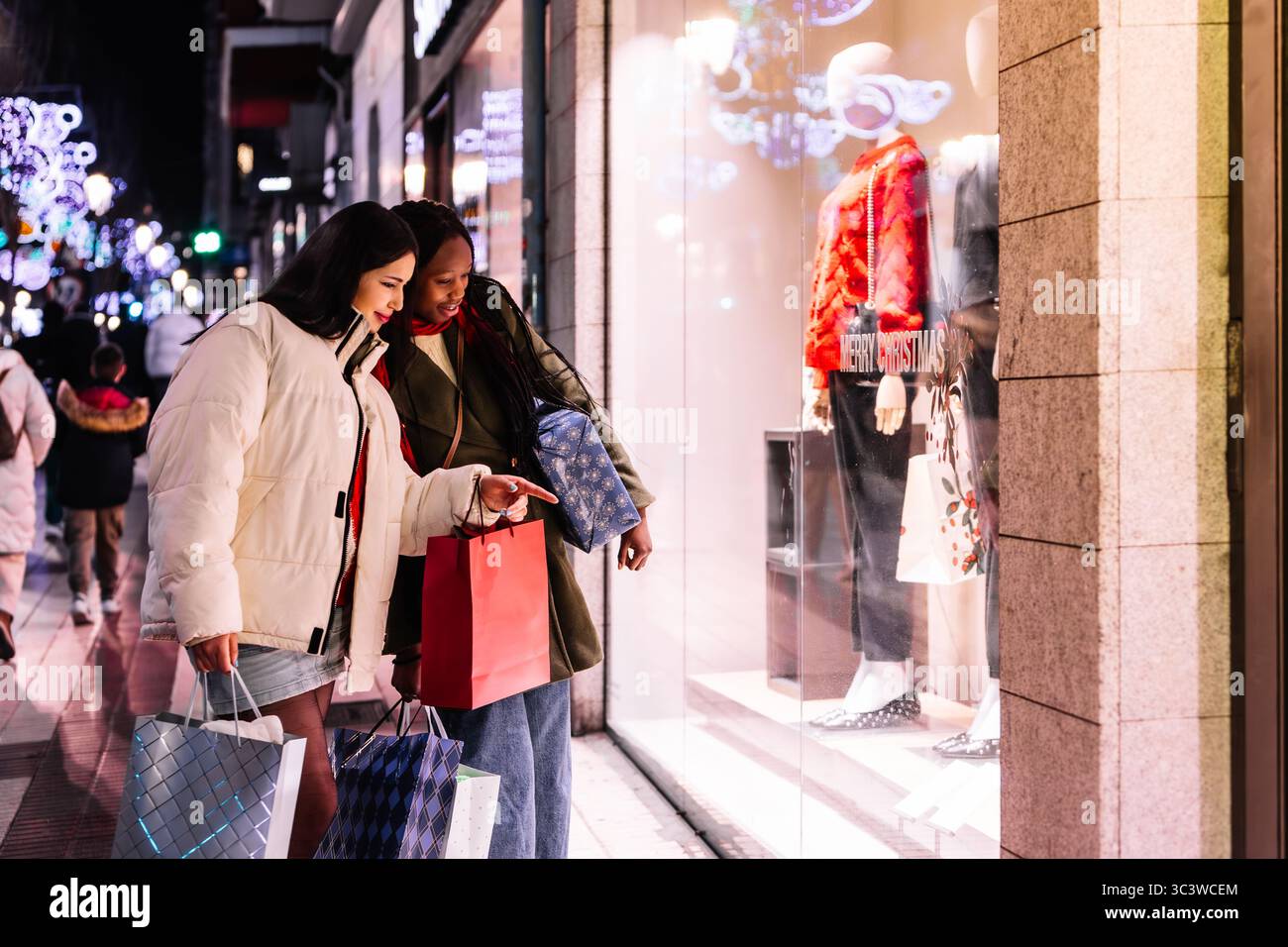 Zwei junge Frauen schauen durch ein festliches Schaufenster mit Schaufensterpuppen in saisonalen Outfits, während sie während eines Abendessens Einkaufstaschen halten Stockfoto