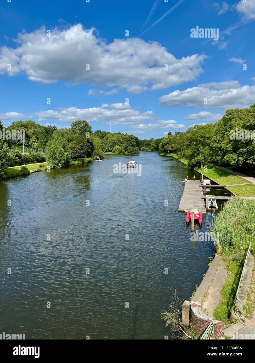 Malerischer Blick auf einen Fluss mit üppigen grünen Bäumen und ein kleines Dock in Lübeck an einem sonnigen Tag. - Smartphone-aufgenommenes Stockfoto