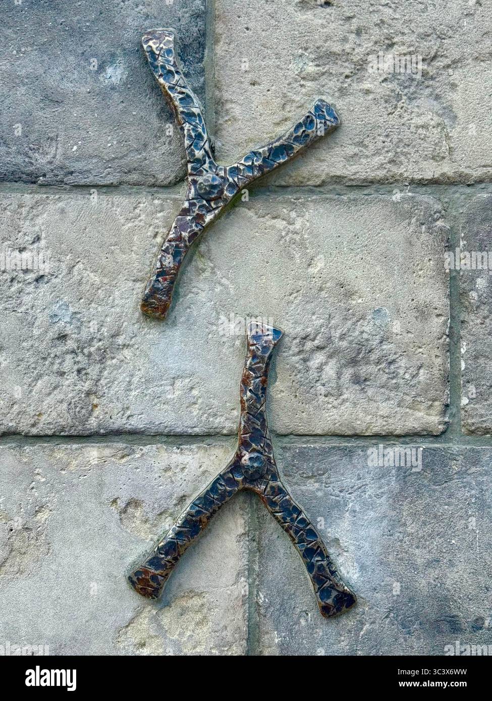 Nahaufnahme zweier dekorativer Y-förmiger Metallzweige, die an einer strukturierten Steinwand befestigt sind. Die metallischen Elemente verfügen über eine gemusterte Oberfläche, die sich im Kontrast zu W - Smartphone-aufgenommenes Stockfoto