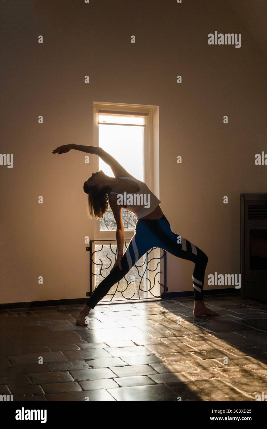 Schlanke, sportliche Frau, die zu Hause vor dem Fenster posiert, wo goldenes Sonnenlicht kommt. Yoga-Routine am Morgen, Selbstpflege, aktiver Lebensstil Stockfoto