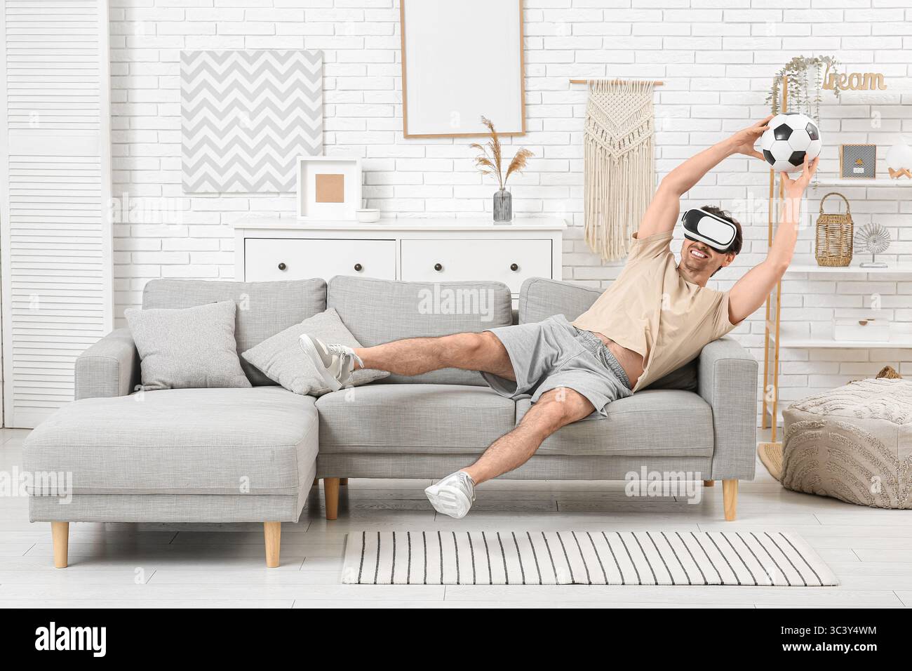 Sportlicher junger Mann in VR-Brille mit Fußball auf Sofa zu Hause Stockfoto Sportlicher junger Mann in VR-Brille mit Fußball auf Sofa zu Hause Stockfoto