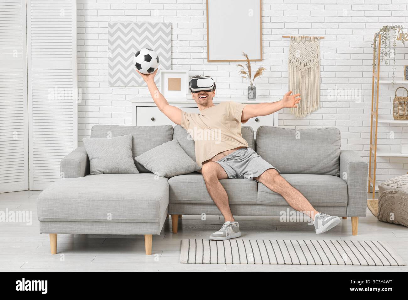 Sportlicher junger Mann in VR-Brille mit Fußball auf Sofa zu Hause Stockfoto Sportlicher junger Mann in VR-Brille mit Fußball auf Sofa zu Hause Stockfoto