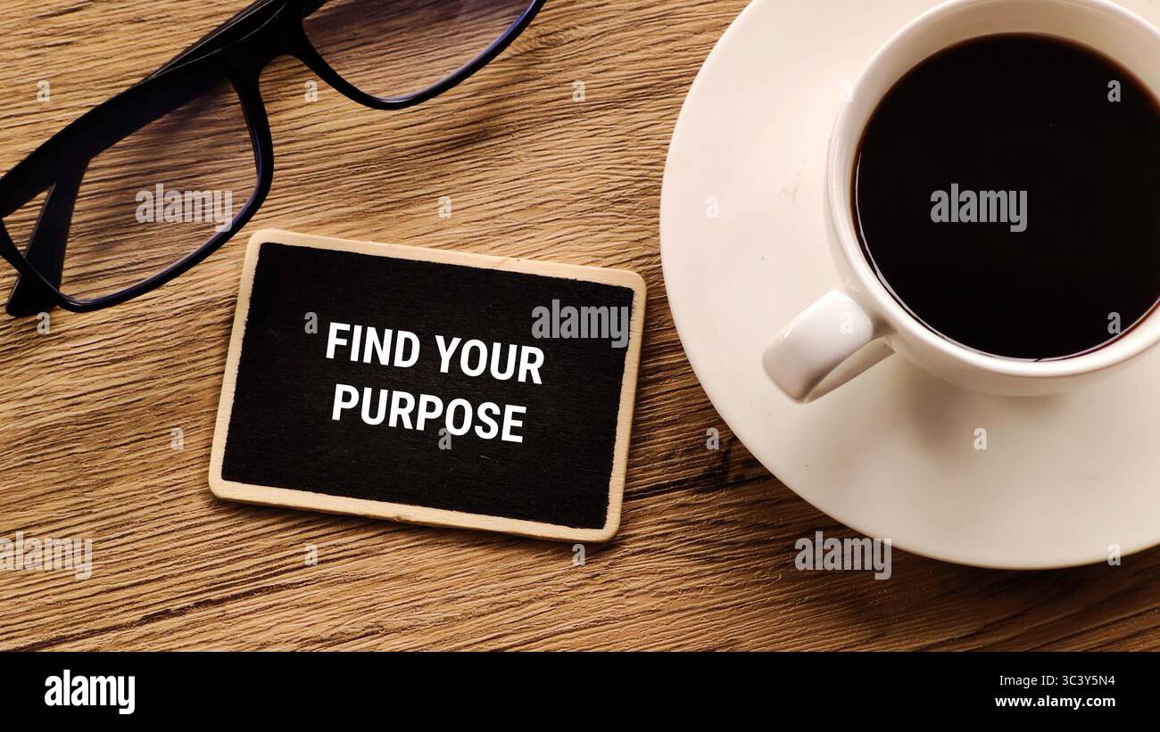Blick von oben auf einen hölzernen Schreibtisch mit einer kleinen Tafel mit der Aufschrift „Find your purpose“, einer Tasse Kaffee und einem Paar Gläser. Stockfoto