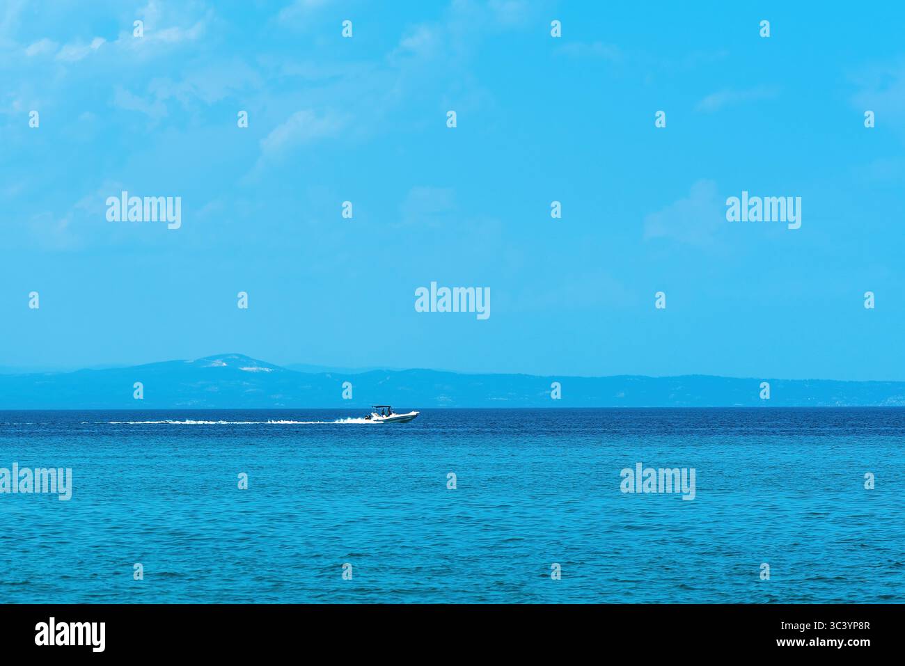 Ein kleines Motorboot fährt über das pulsierende blaue Meer und hinterlässt einen weißen Pfad. In der Ferne dehnen sich schwache Konturen hügeligen Geländes acro Stockfoto