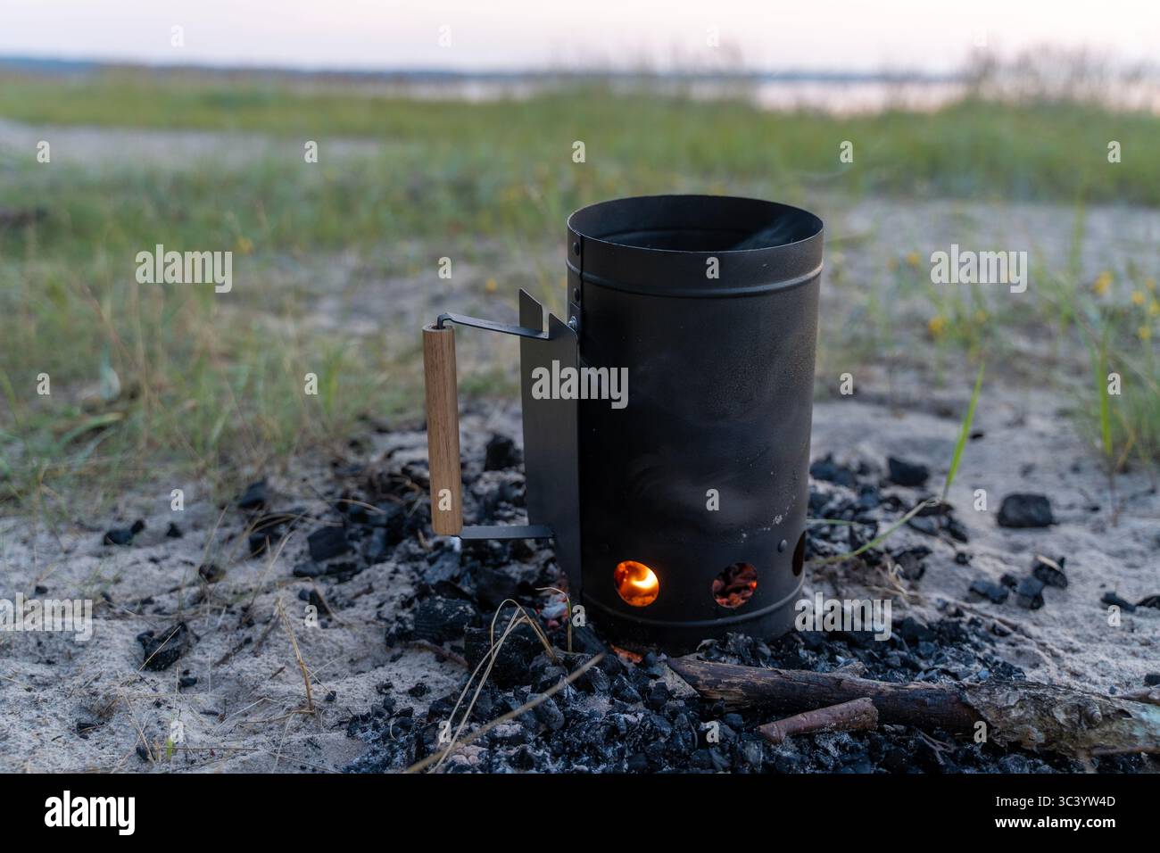 Holzkohle-Schornsteinstarter mit glühenden Kohlen am Sandstrand - umweltfreundliches Feuerbeleuchtungswerkzeug für Grill und Camping-Kochen im Freien Stockfoto