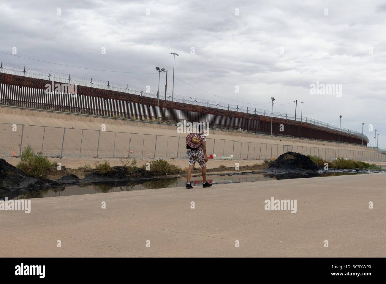 Eine Einzelfigur navigiert einen Pfad am Grenzzaun Ciudad Juarez, einem ergreifenden Symbol für Migrationsherausforderungen unter einem bewölkten Himmel. Stockfoto