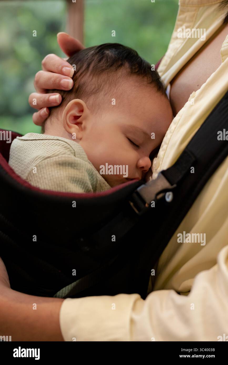 Pflegerin, die die Kleinkind-Tochter in Strickjacke in Fronttrage zu Hause am Fenster schläft Stockfoto