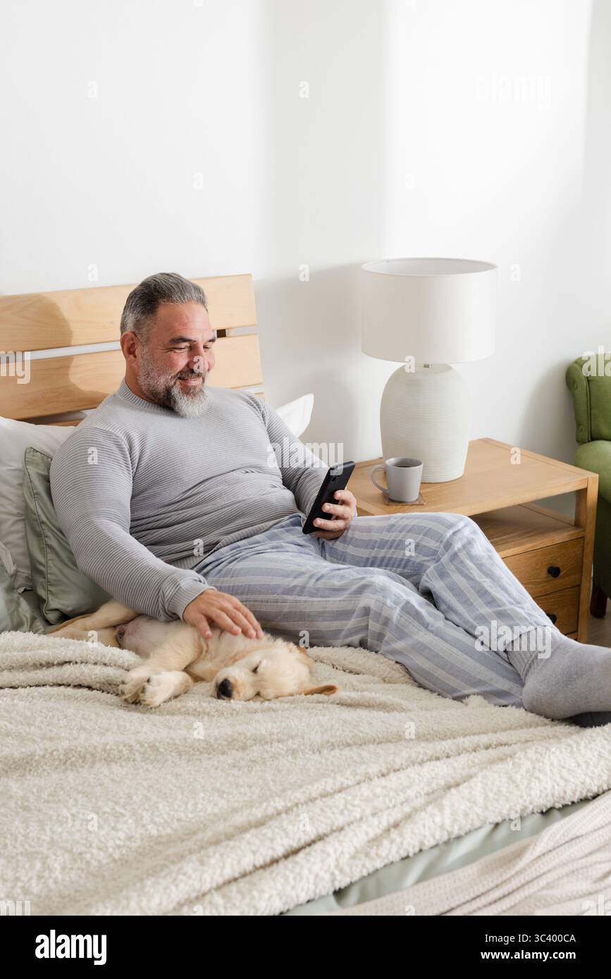 Ein älterer Mann, der auf dem Bett sitzt, einen Pyjama trägt, das Smartphone hält und den Welpen unter der Decke streichelt Stockfoto