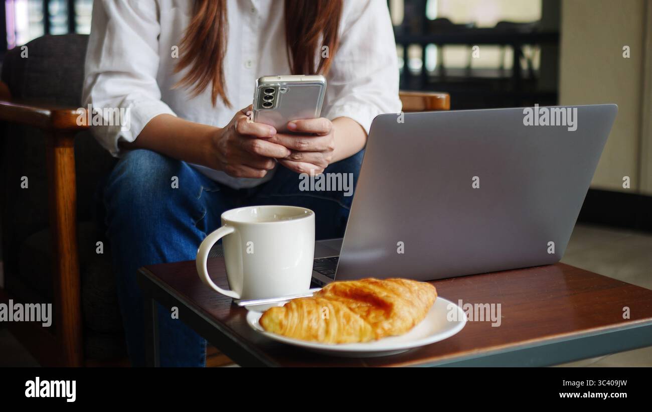 Vorderansicht einer Frau, die zu Hause Telefon und Laptop benutzt oder im Café für die Arbeit am Morgen mit heißem Kaffee, anonymes Gesicht. Arbeiten von zu Hause aus Stockfoto