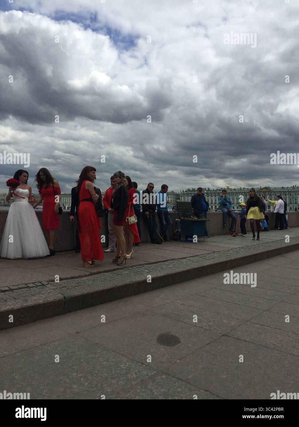 Eine Gruppe von Menschen, darunter eine Braut in weiß und andere in roten Kleidern, versammeln sich auf einer Uferpromenade in Sankt Petersburg unter einem bewölkten Himmel Stockfoto