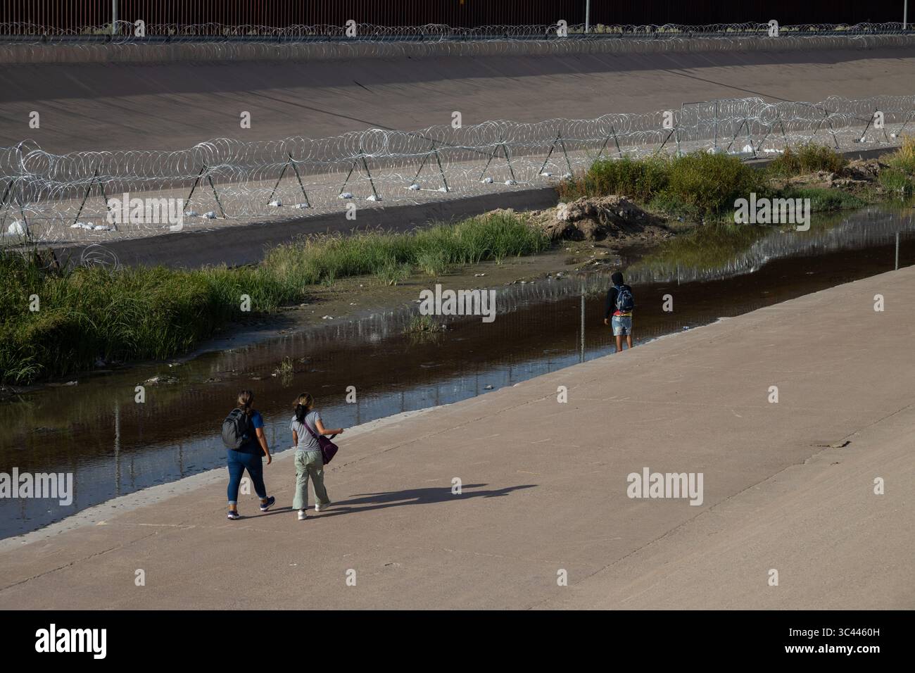 Drei Personen gehen an einem Kanal in der Nähe von Ciudad Juarez entlang, flankiert von Stacheldraht, was die Herausforderungen der Migration verdeutlicht. Stockfoto