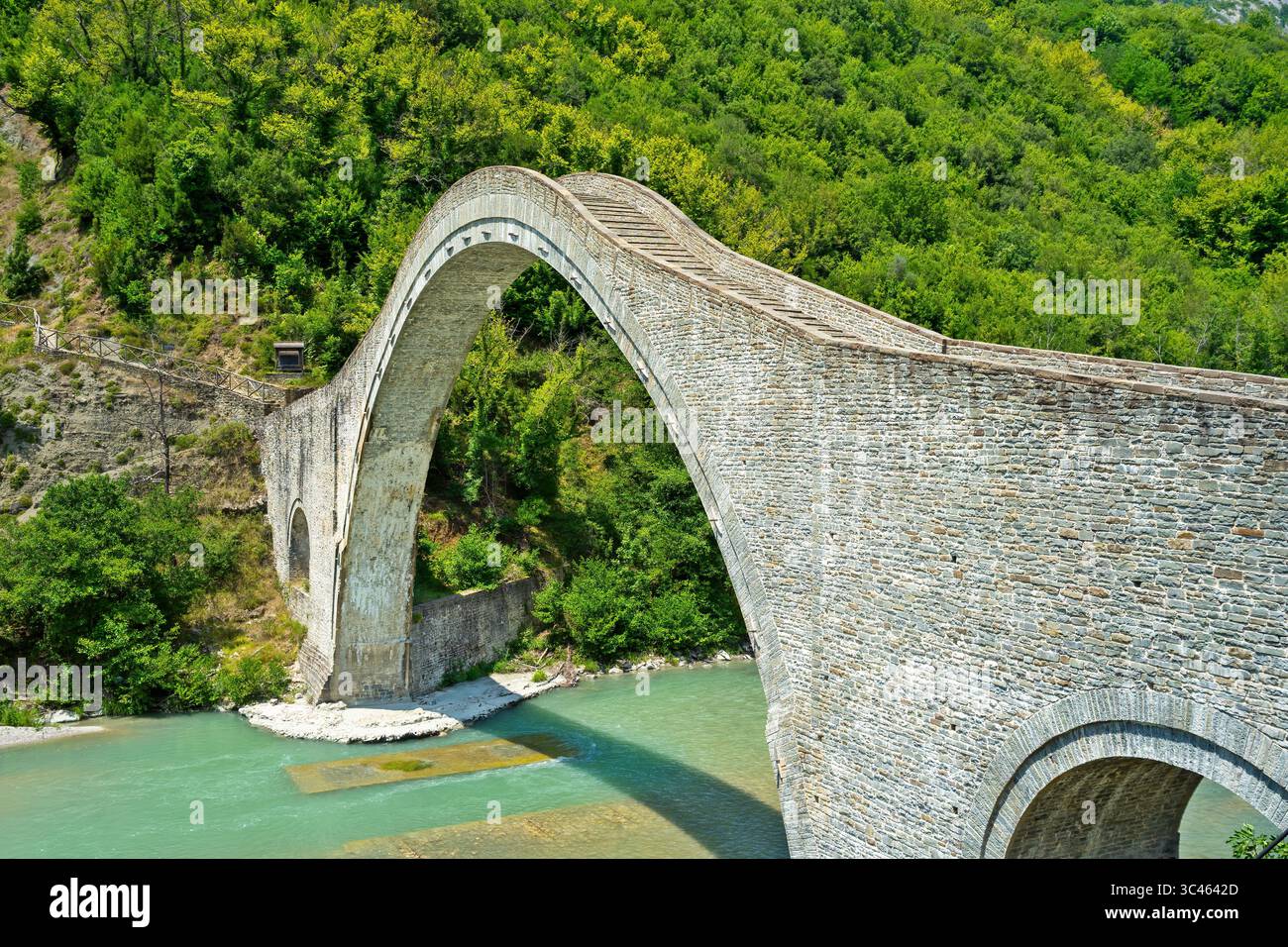 Die historische Plaka-Brücke überspannt den Arachthos-Fluss in Epirus, Griechenland, umgeben von üppig grünen Wäldern und kristallklarem Wasser an einem hellen Sommertag Stockfoto