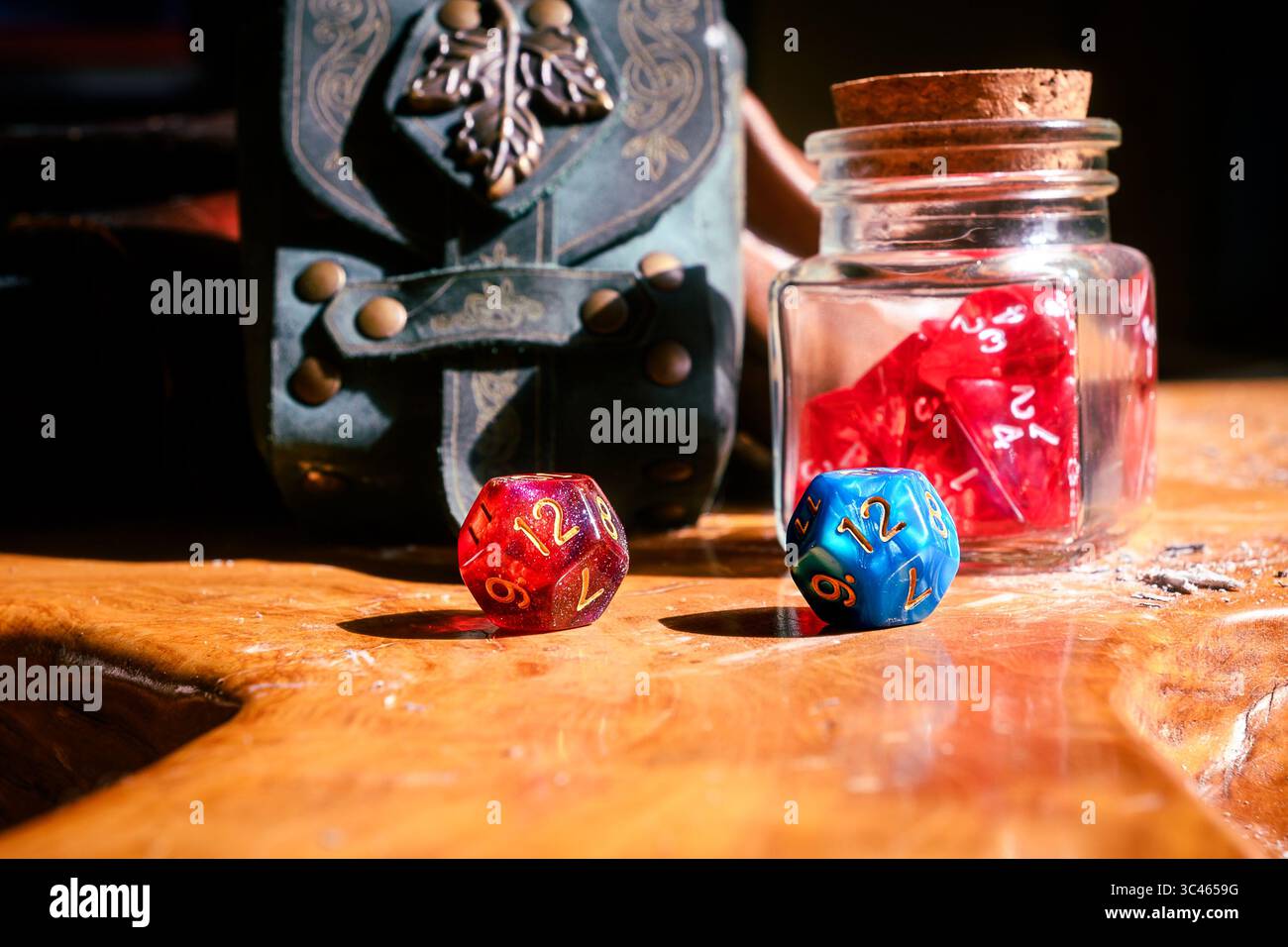Eine rot-blaue D12-Stanzform ruht auf einer Holzoberfläche neben einem Würfelbecher und einer dekorativen Ledertasche. Stockfoto