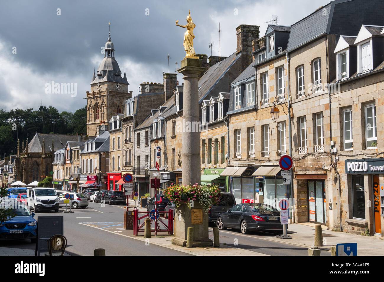 Villedieu-les-Poêles, Frankreich - 21. juli 2025: Blick von der Hauptstraße mit Glockenturm der Kirche Notre-Dame und Marianne-Statue unter stürmischem Himmel Stockfoto