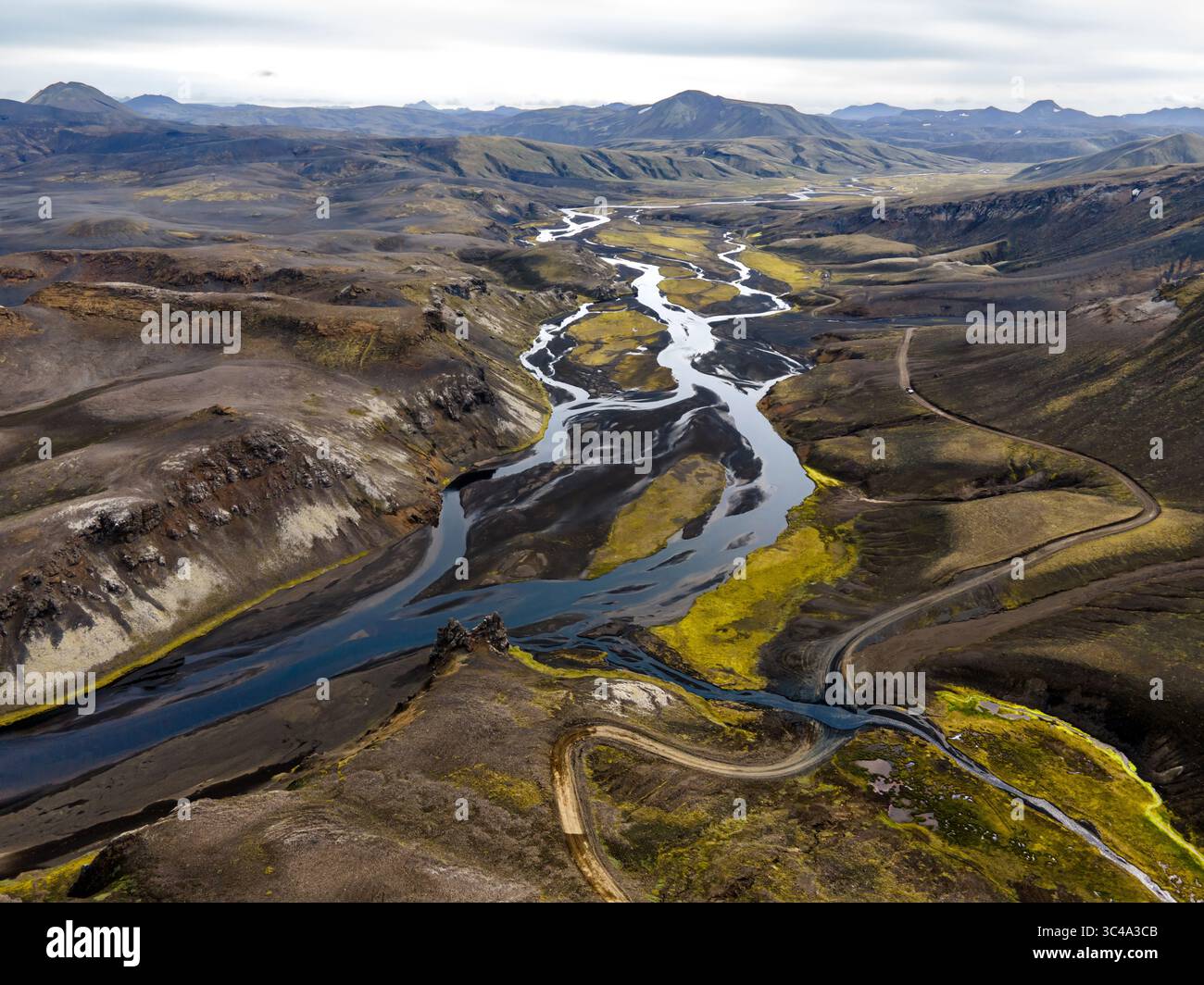 Aus der Vogelperspektive auf einen geflochtenen Fluss, der sich durch das Hochland schlängelt, mit einem Kontrast zu schwarzem Sand und leuchtendem grünem Moos, einer abgelegenen Straße, die ihren Weg entlang zieht, Kirkjubaejarklaustur, Island. Stockfoto