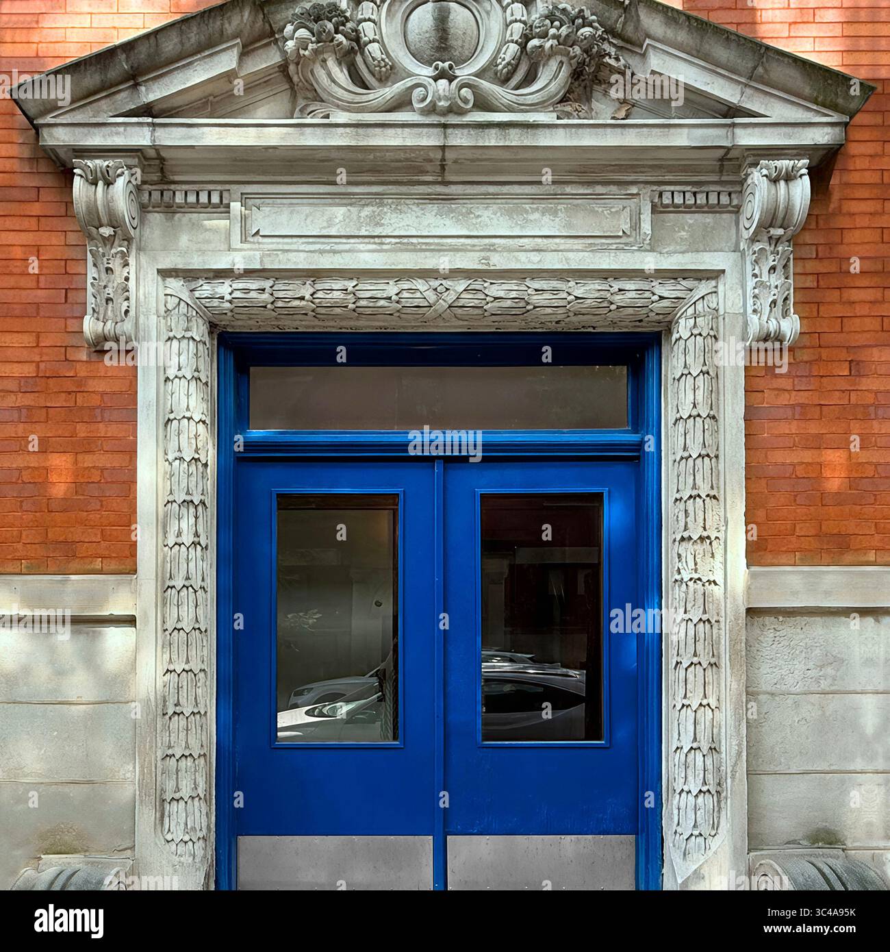 Die doppelten Türen in leuchtend blauem Blau, eingerahmt von geschnitztem Mauerwerk und einem dekorativen Giebel, befinden sich in einer roten Backsteinfassade eines historischen Gebäudes im Beaux-Arts-Stil. - Smartphone-aufgenommenes Stockfoto