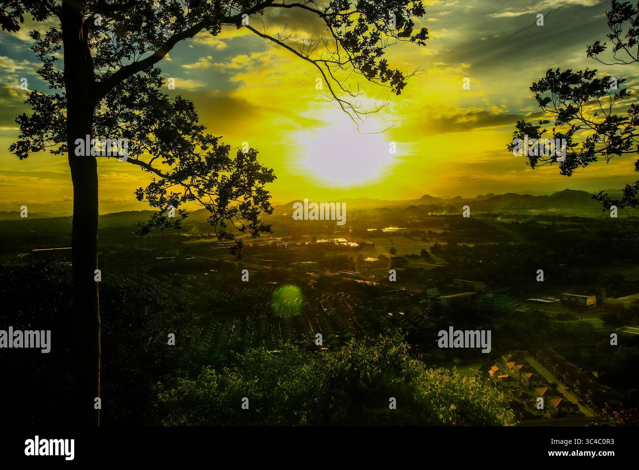 Malerischer Blick in die Dämmerung vom Berggipfel aus mit Blick auf das Dorf, Reisfelder und tropischen Wald unter einem leuchtenden goldenen Himmel. Silhouettenbäume im Vordergrund. Stockfoto