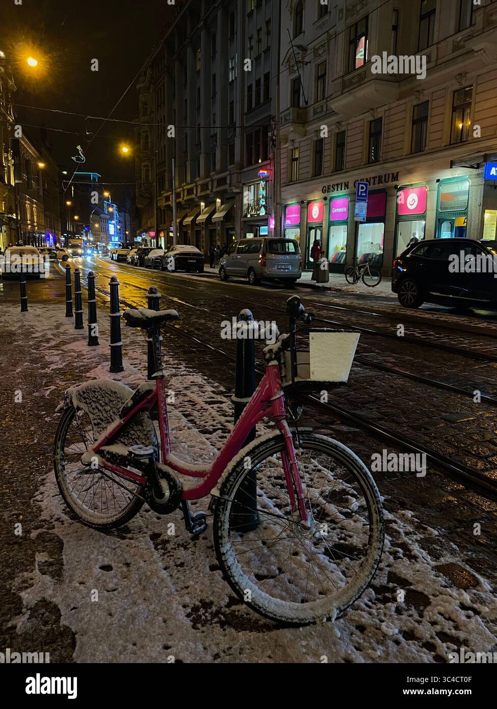 Schneebedecktes Fahrrad parkt in einer ruhigen Straße in Prag, Tschechien, in einer verschneiten Winternacht mit leuchtenden Straßenlampen. - Smartphone-aufgenommenes Stockfoto