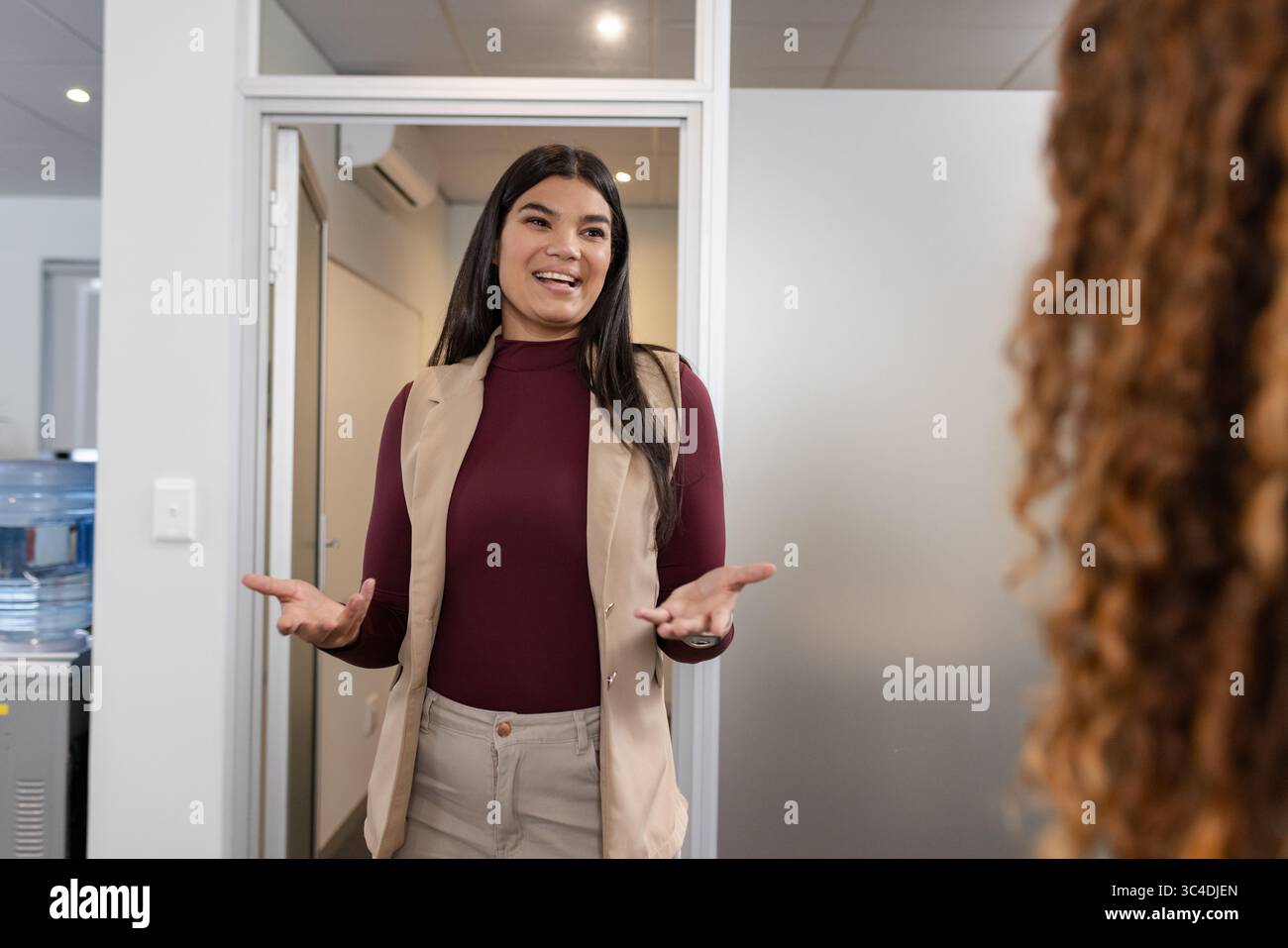 Verschiedene weibliche Kollegen, die im Flur des Büros stehen, tragen Blazer, Gesten in der Nähe des Wasserkühlers Stockfoto