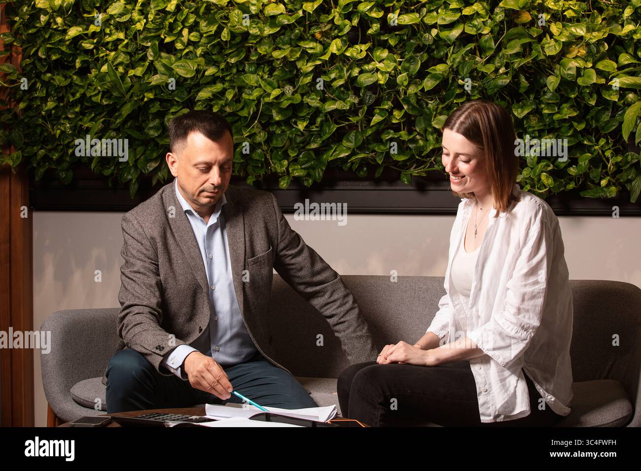 Zwei Geschäftspartner lachen zusammen, während sie in einem modernen Büro über Dokumente diskutieren. Freundliche Atmosphäre, grüne Pflanzenwand, Teamwork. Stockfoto