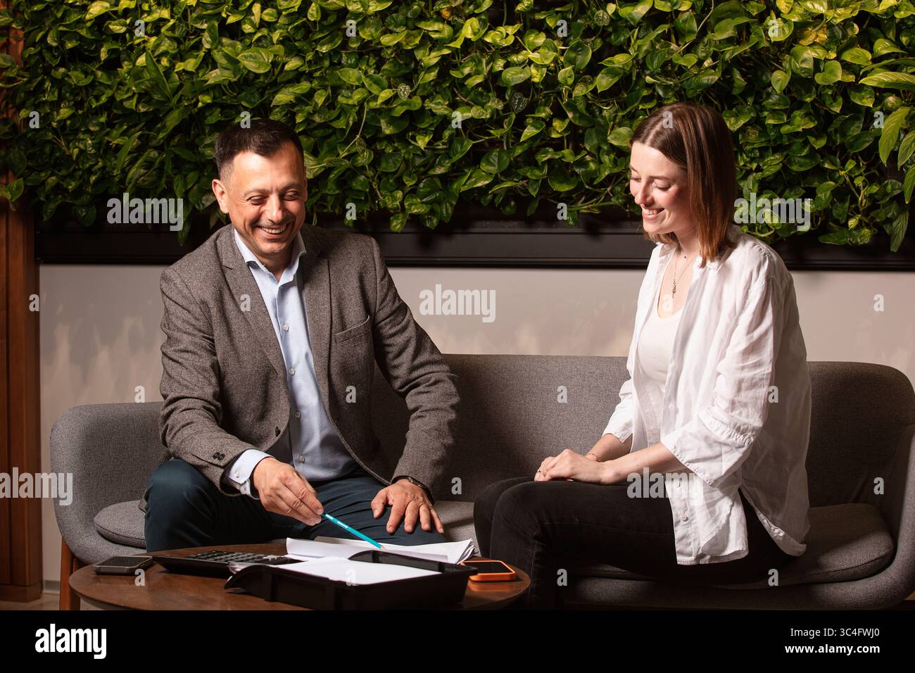 Zwei Geschäftspartner lachen zusammen, während sie in einem modernen Büro über Dokumente diskutieren. Freundliche Atmosphäre, grüne Pflanzenwand, Teamwork. Stockfoto