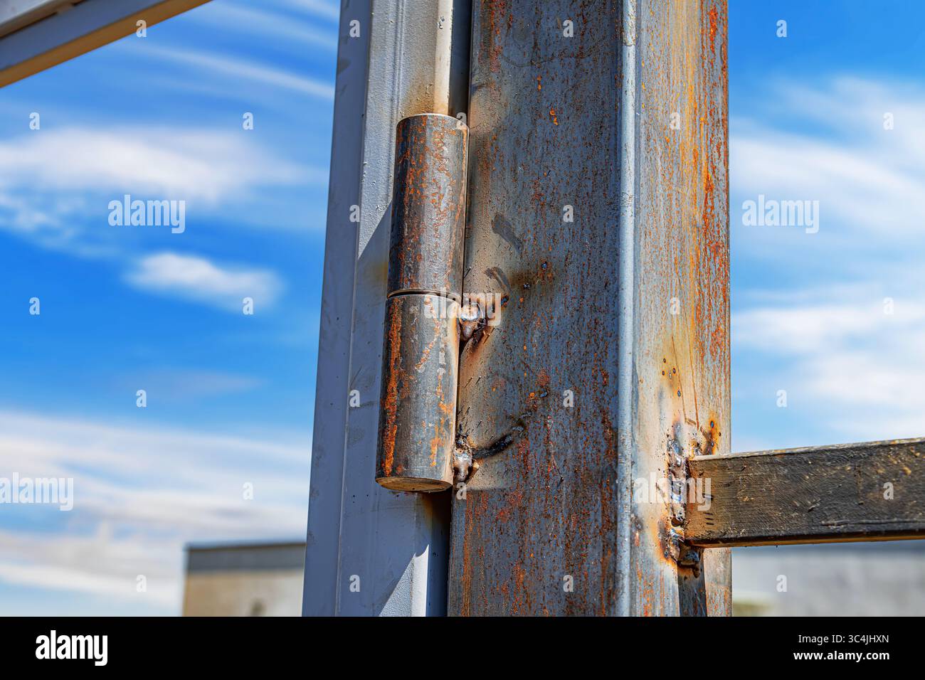 Ein rostiges Metallscharnier, das unter einem hellblauen Himmel an eine Stahlrahmenkonstruktion geschweißt ist und Anzeichen von Witterung und Korrosion zeigt. Stockfoto
