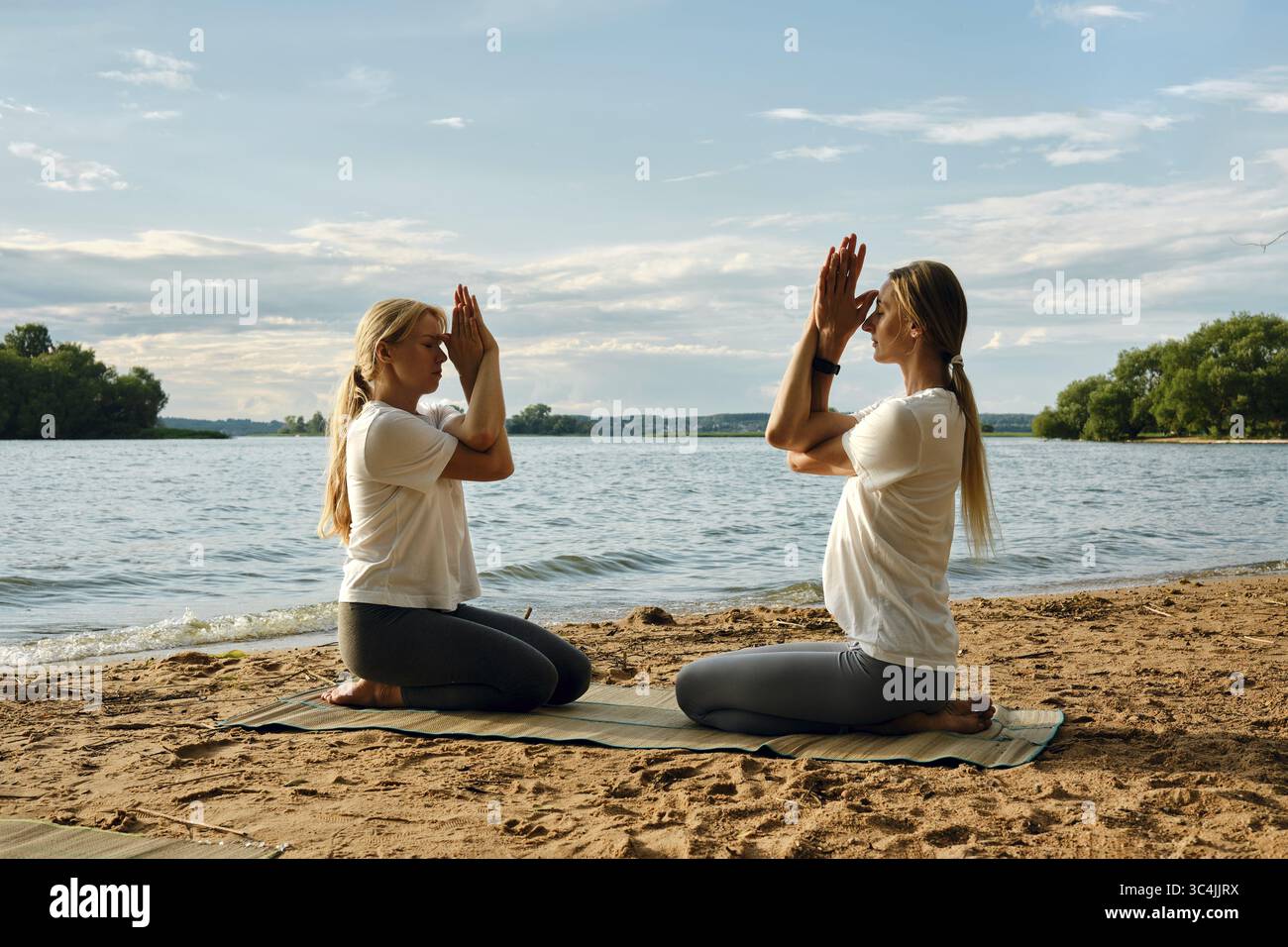 Zwei Personen nehmen an einer Yoga-Sitzung an einem Sandstrand an einem ruhigen See Teil. Die Sonne geht unter und wirft sanfte Farben über den Himmel. Sie führen ein Ich aus Stockfoto