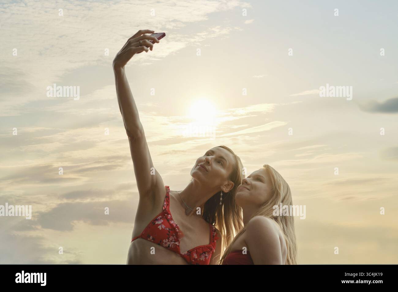 Zwei Freunde fangen einen freudigen Moment ein, indem sie ein Selfie mit dem Sonnenuntergang hinter ihnen machen. In Bikinis gekleidet machen sie Fotos, um sich an ihren Urlaub zu erinnern Stockfoto Zwei Freunde fangen einen freudigen Moment ein, indem sie ein Selfie mit dem Sonnenuntergang hinter ihnen machen. In Bikinis gekleidet machen sie Fotos, um sich an ihren Urlaub zu erinnern Stockfoto