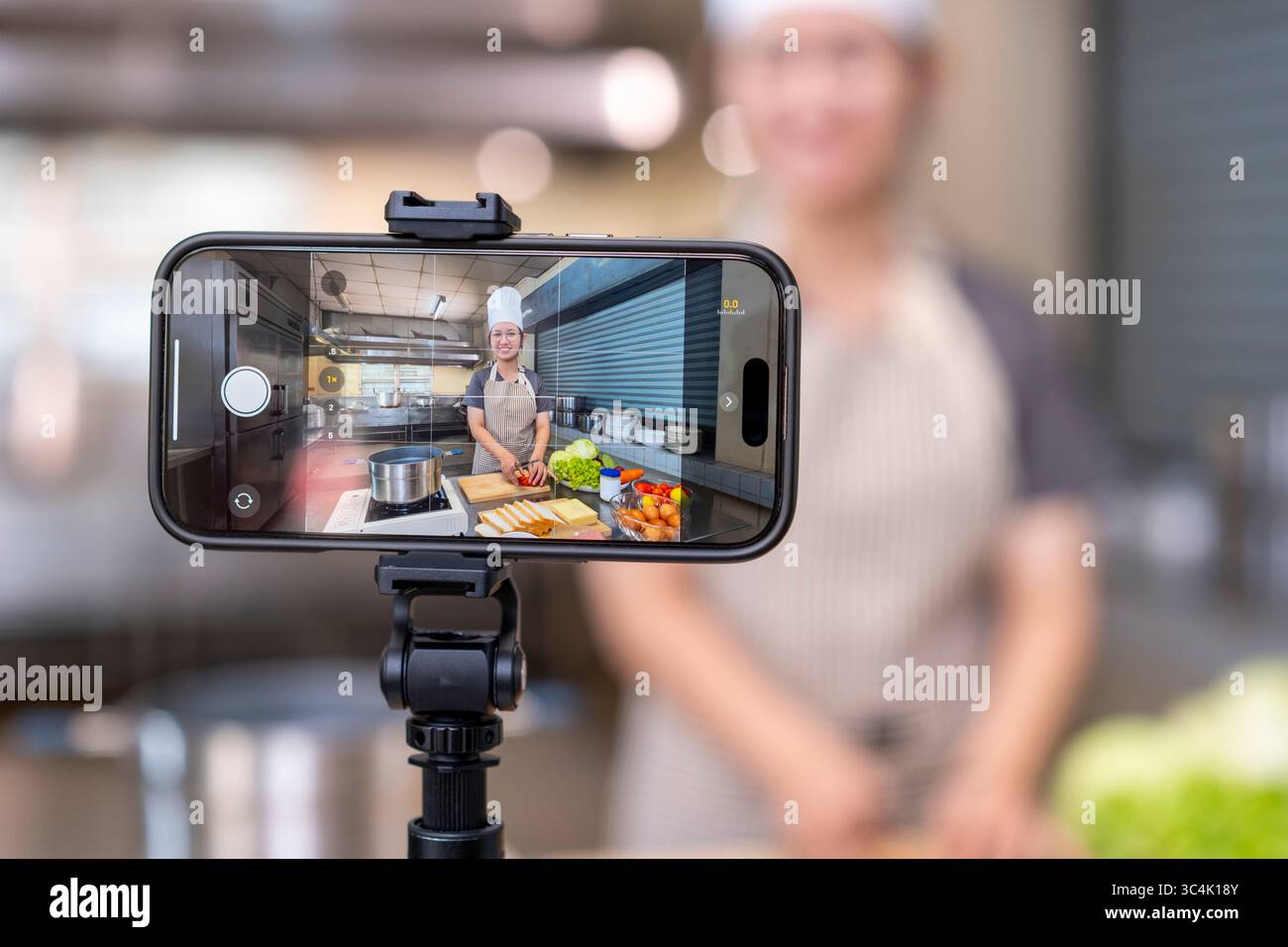 Ein Smartphone auf einem Stativ nimmt eine lächelnde junge Erwachsene asiatische Chefin auf, die in einer professionellen Küche Essen zubereitet. Perfekt für Online-Kochkurse Stockfoto