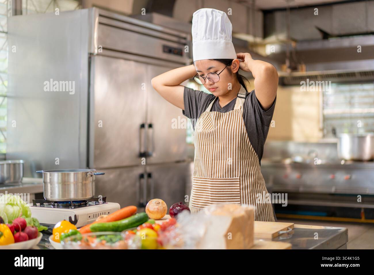 Eine junge asiatische Küchenchefin, die Brille und eine gestreifte Schürze trägt, passt ihre traditionelle weiße Kochmütze in einer professionellen Küche an. Idee Stockfoto