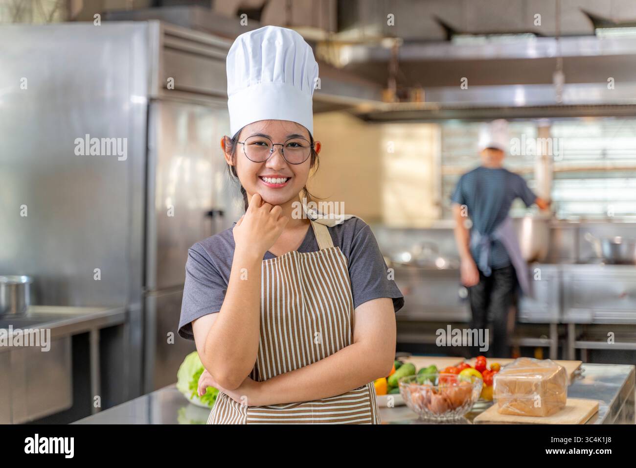 Eine glückliche junge Erwachsene asiatische Chefin mit überkreuzten Armen lächelt selbstbewusst in einer geschäftigen professionellen Küche mit einem anderen Koch im Hintergrund. Ide Stockfoto