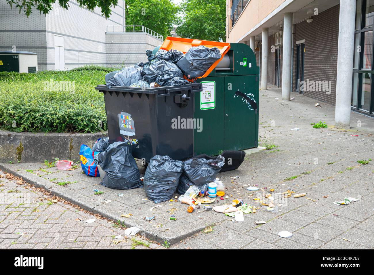 Müll in einem überfüllten Mülltonne vor einem Wohngebäude, umgeben von Müll auf dem Boden. Stockfoto