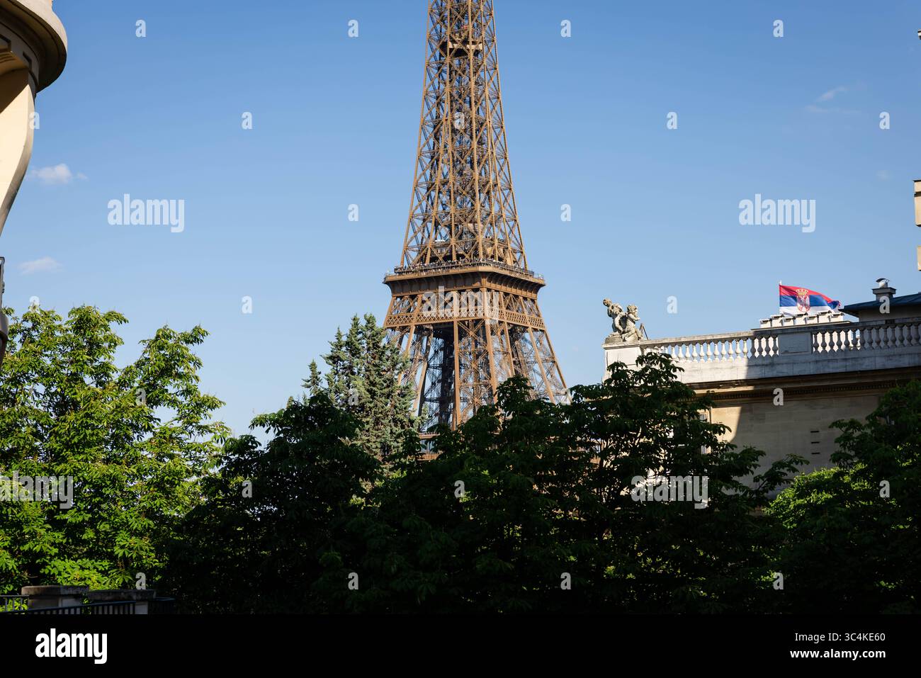 Ein majestätischer Eiffelturm, teilweise von üppigen grünen Bäumen verdeckt, unter einem leuchtend blauen Himmel. Ein Gebäude mit serbischer Flagge fügt Intrigen hinzu. Ideal für TRA Stockfoto