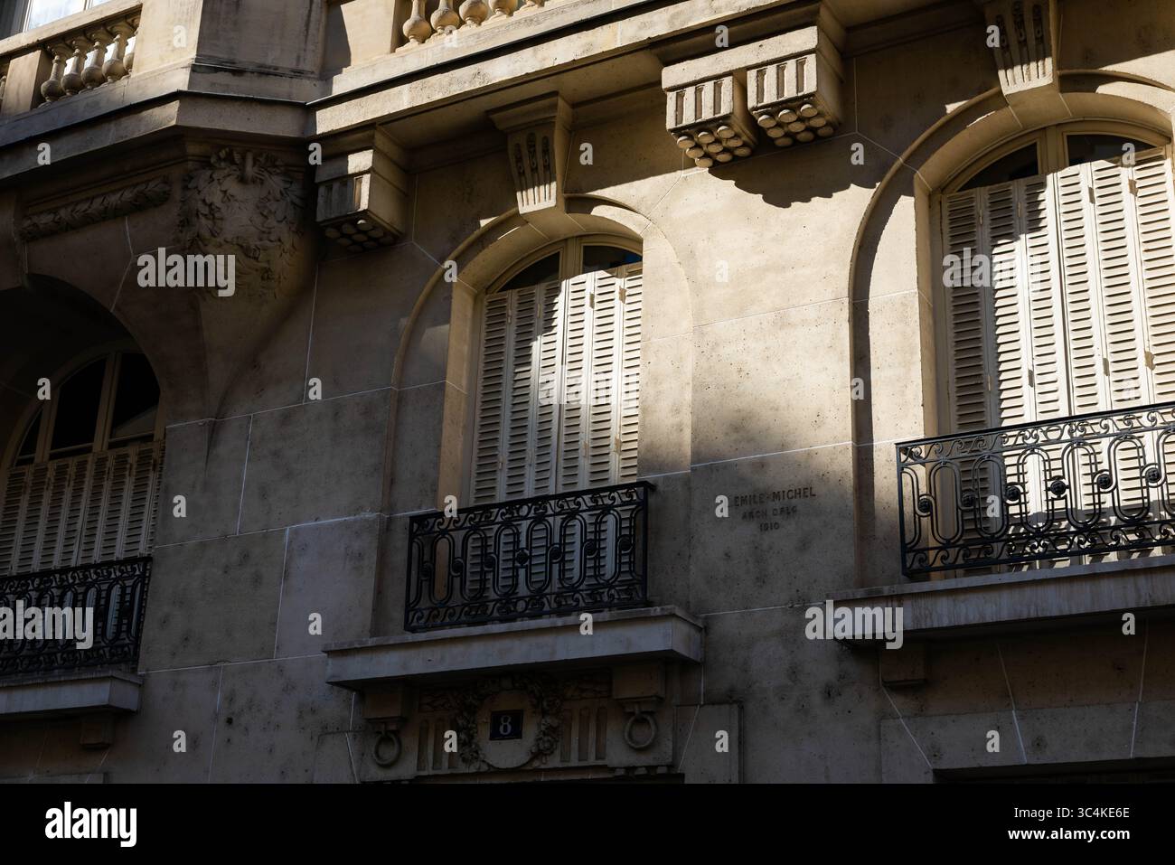 Sonnenverwöhnte Pariser Gebäudefassade; Bogenfenster mit weißen Fensterläden und kunstvoll verzierte eiserne Balkone. Elegante architektonische Details und dezente Schatten Stockfoto