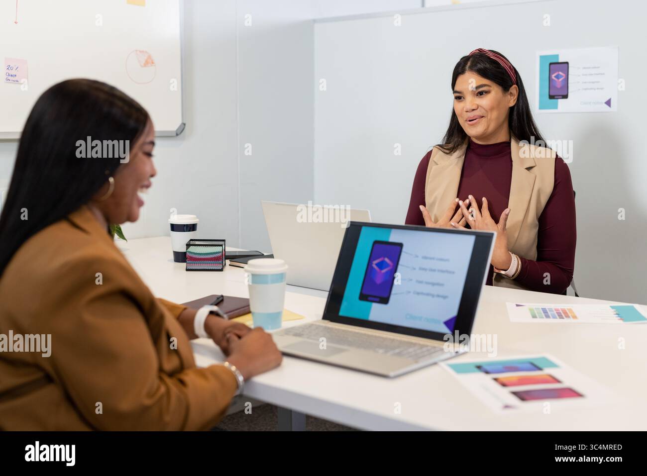 Verschiedene weibliche Kollegen diskutieren im Besprechungsraum mit Laptops und Mockups über das Design mobiler Apps Stockfoto