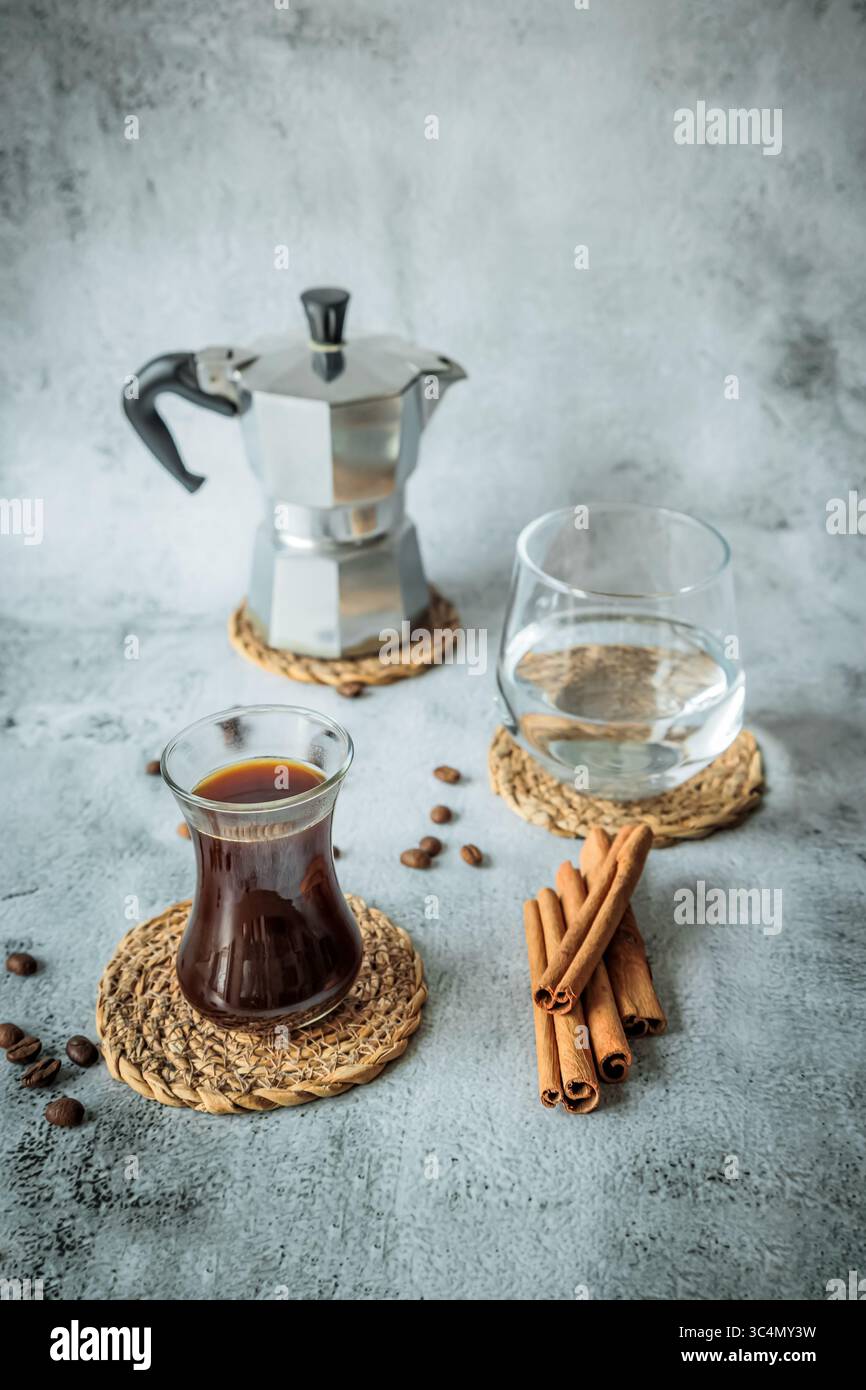 Schwarzer Kaffee in geschwungenem Glas mit Wasserbecher, Mokakanne und Zimtstangen auf gewebten Untersetzern, verstreute Bohnen auf hellem Steinhintergrund, luftig vertikal Stockfoto